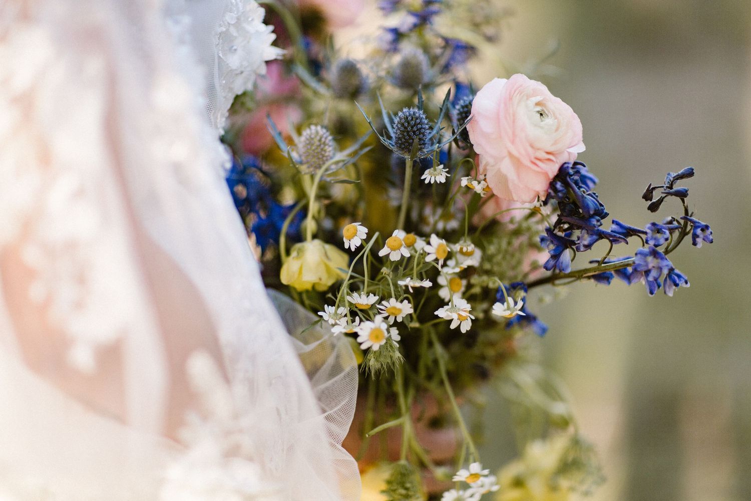 Close-up of wildflower bridal bouquet featuring pink ranunculus, blue thistles, and white daisies.