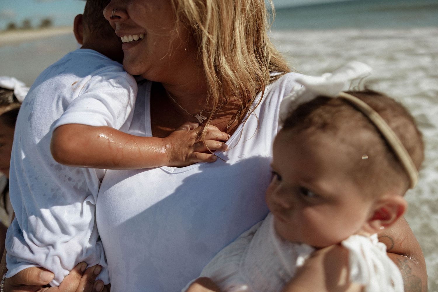 Smiling blonde woman holding two young children on a bright sandy beach.