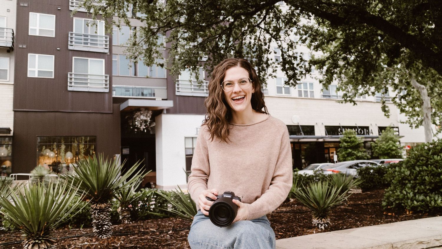 Smiling woman photographer holding camera outdoors near modern building with lush trees.