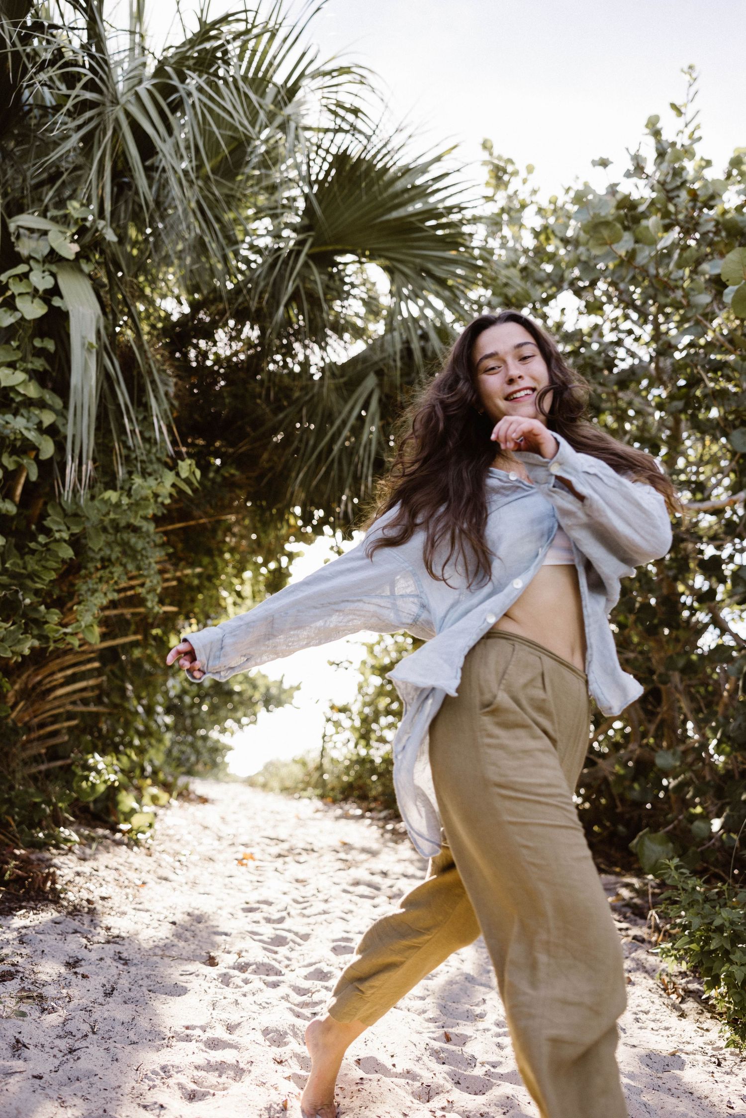 Happy young woman in olive pants and white shirt dancing barefoot on a sandy tropical beach path.