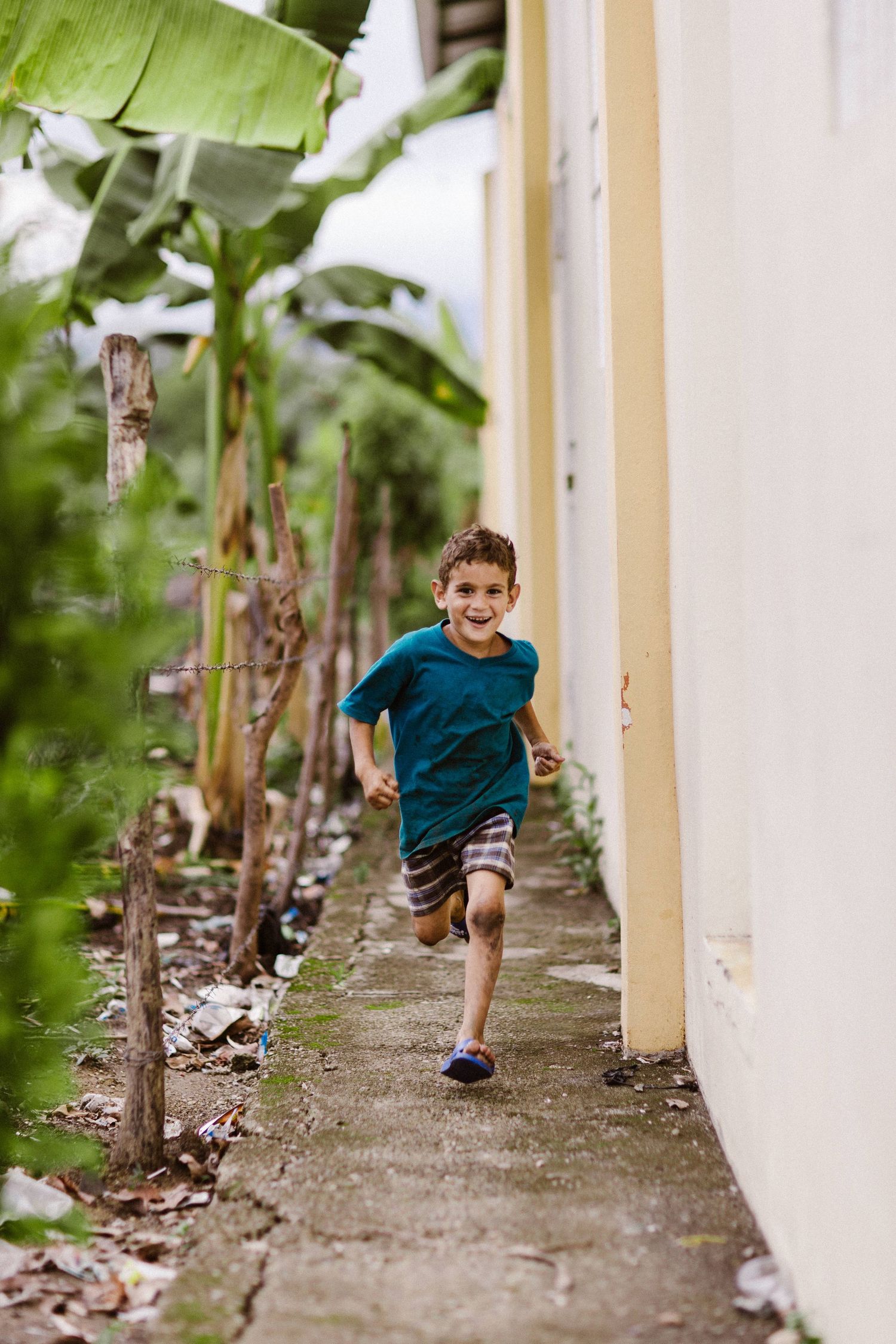A smiling boy in a teal shirt runs along a narrow path beside banana trees and a yellow wall.