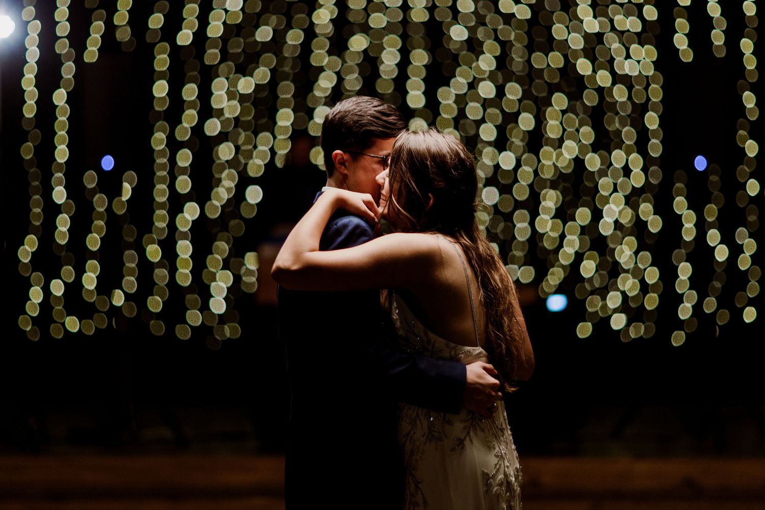 Couple sharing a romantic slow dance embrace surrounded by glowing bokeh lights at a wedding reception.