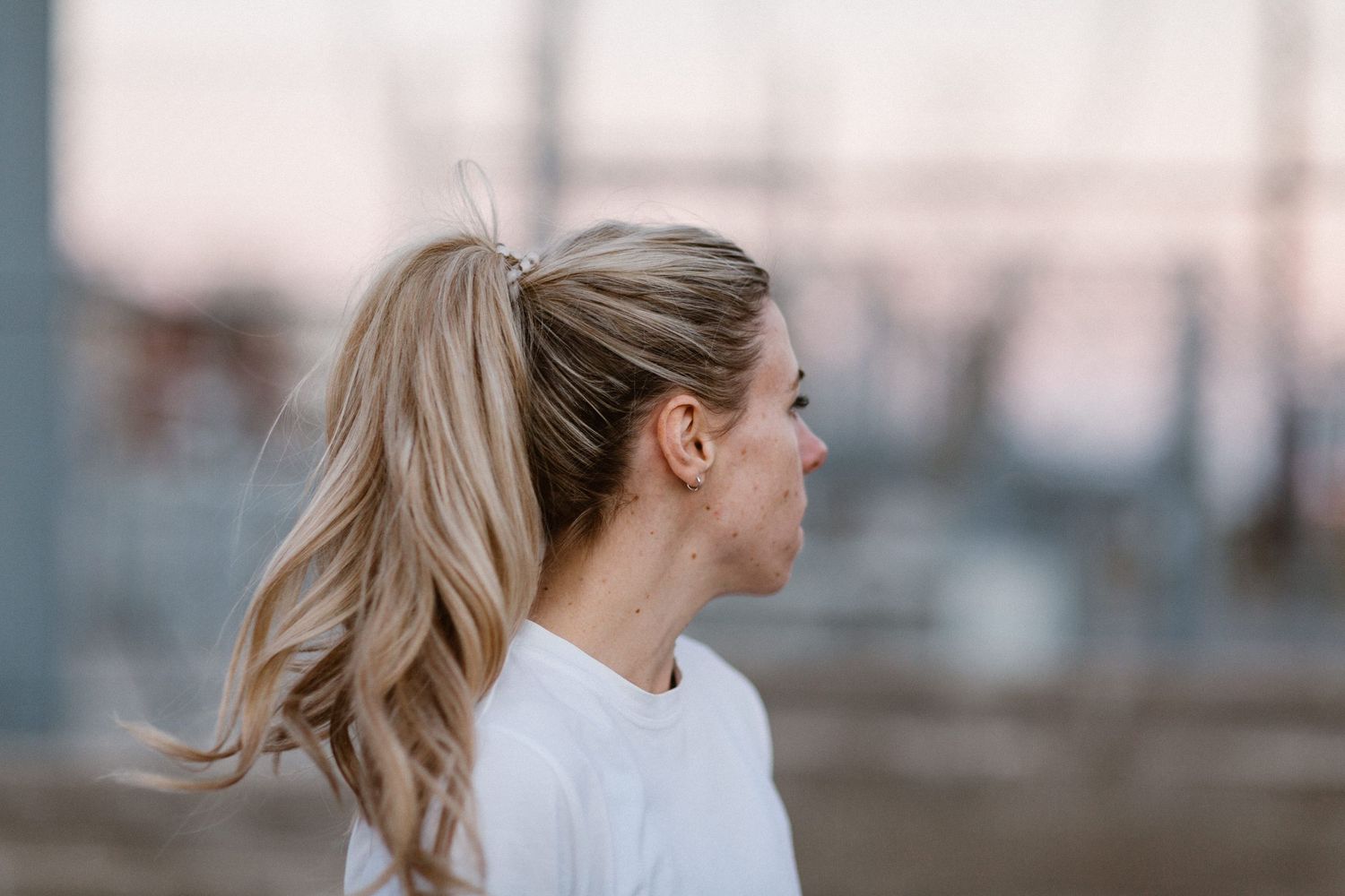 Blonde woman with ponytail wearing white top, photographed in profile against a blurred outdoor background.
