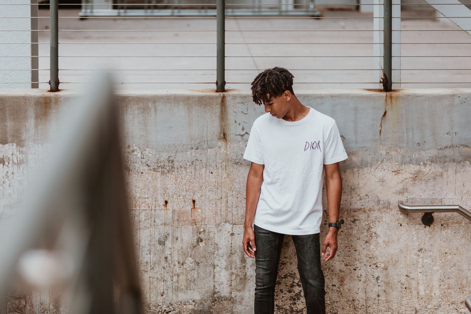 Young man with dreadlocks wearing a white t-shirt and gray jeans standing against a concrete wall outdoors.