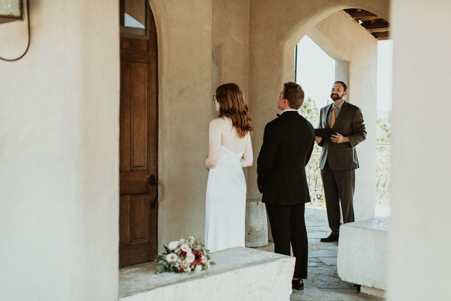 Couple exchanging wedding vows with officiant outdoors under arched venue entrance with floral bouquet nearby.