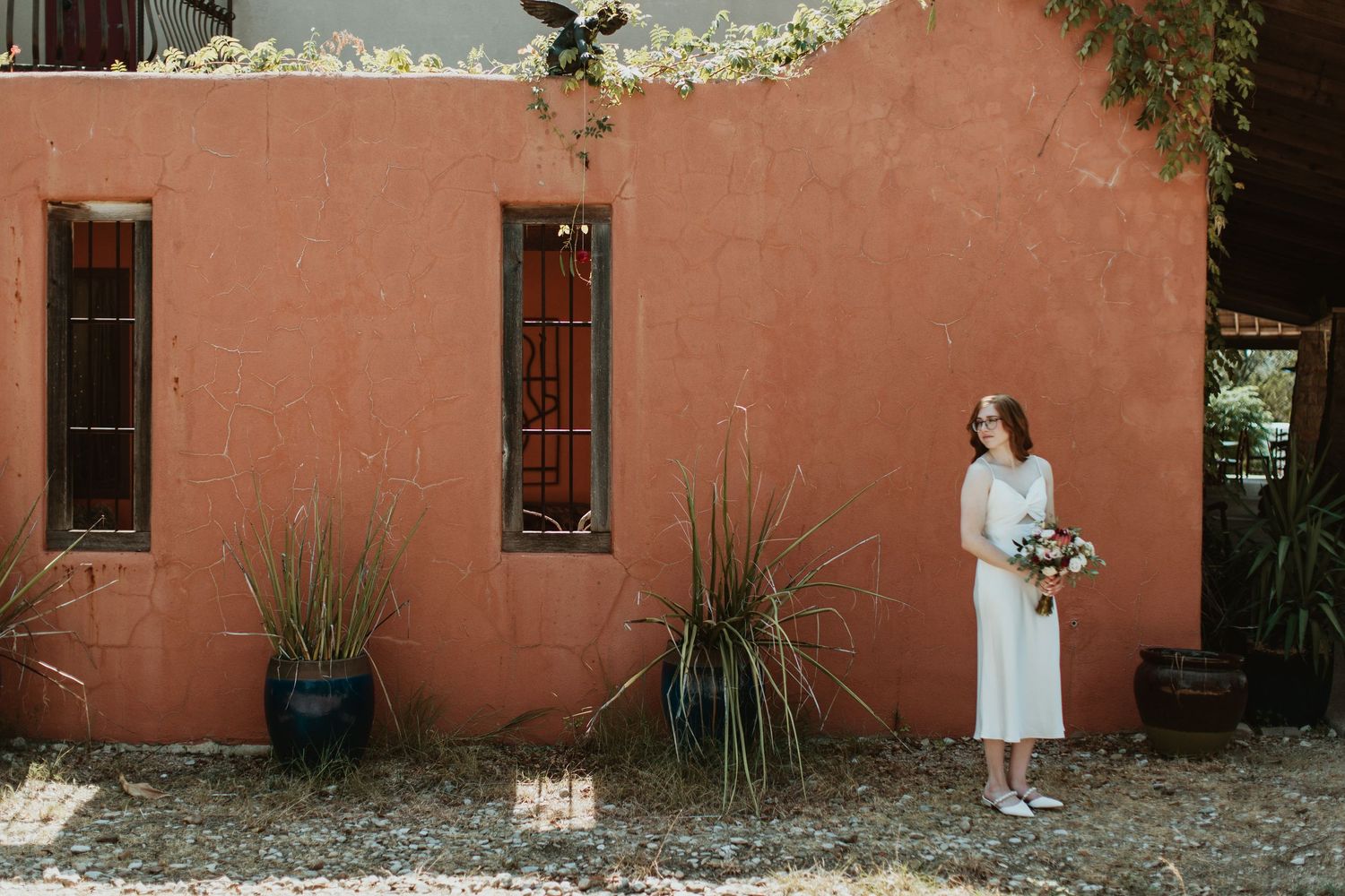 Bride in white dress holding bouquet stands before a rustic terracotta red building with desert plants.