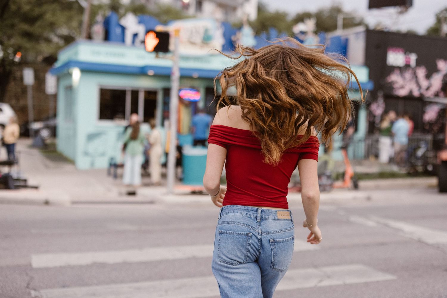 Woman in red off-shoulder top and jeans walking across urban street crosswalk.