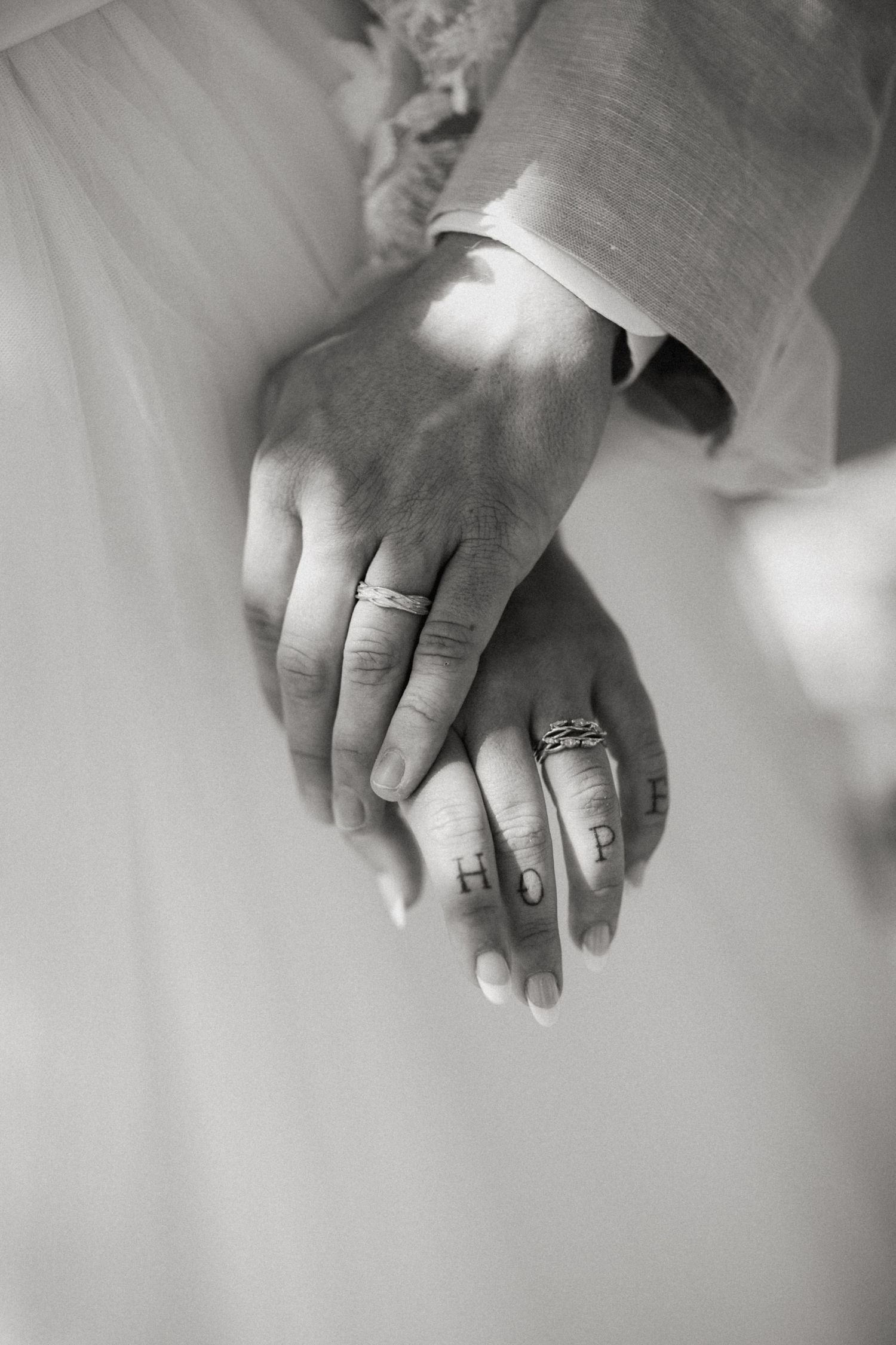 Close-up black and white photo of newlyweds holding hands showing wedding rings and HOPE tattoo.