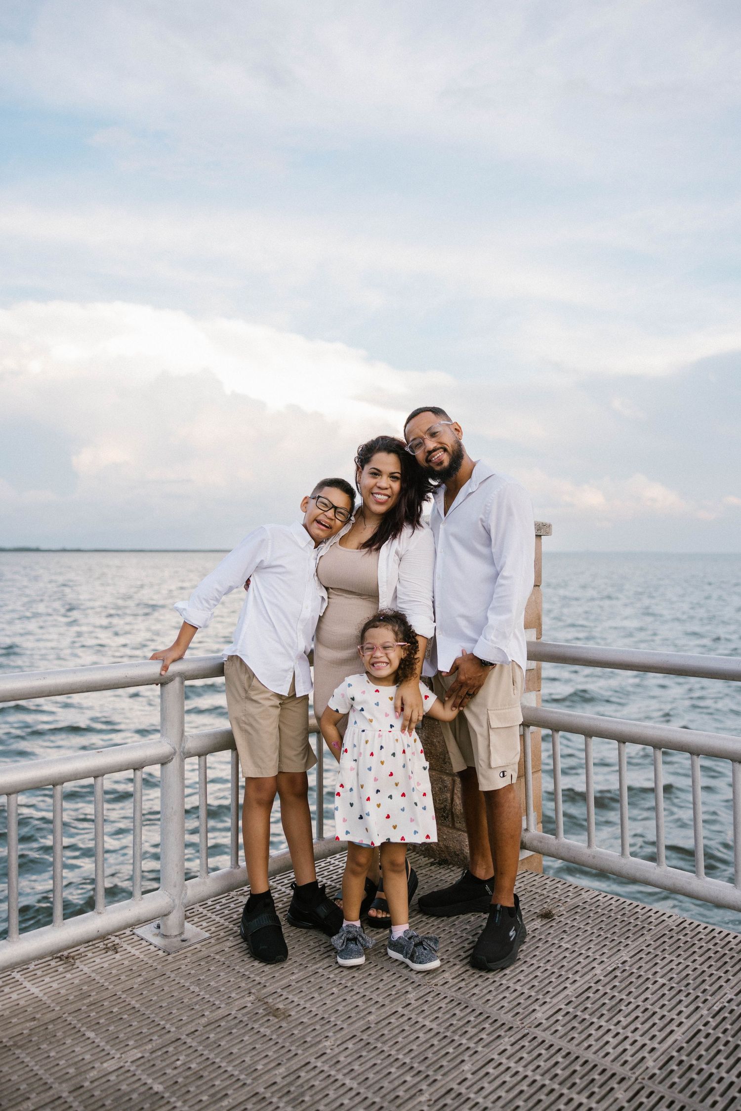 Family of four posing together on a waterfront pier with ocean views and cloudy sky behind them.