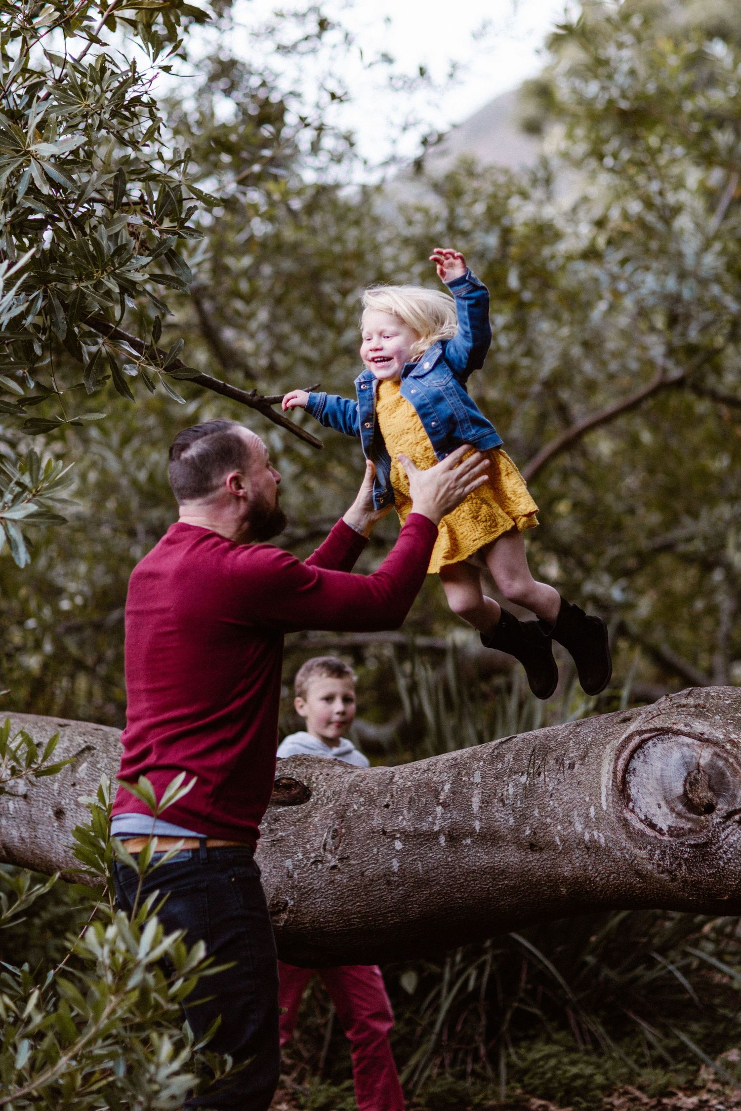 Father lifting laughing blonde girl off a fallen tree trunk in a forest setting.