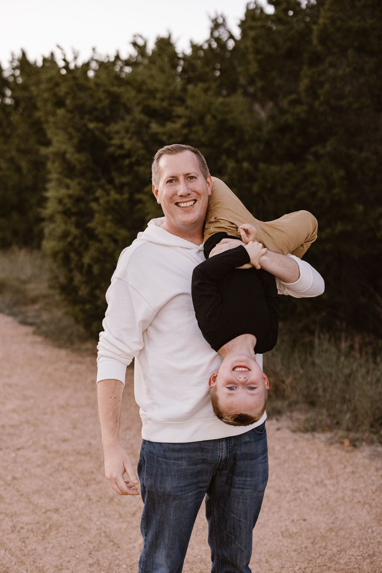 Happy father playfully holding his young child upside down on a sandy trail with evergreen trees behind.
