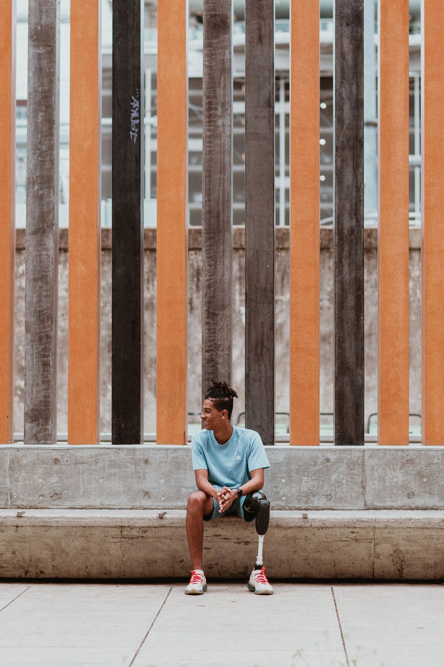 Young man with prosthetic leg sitting on urban bench against colorful wooden slat wall.