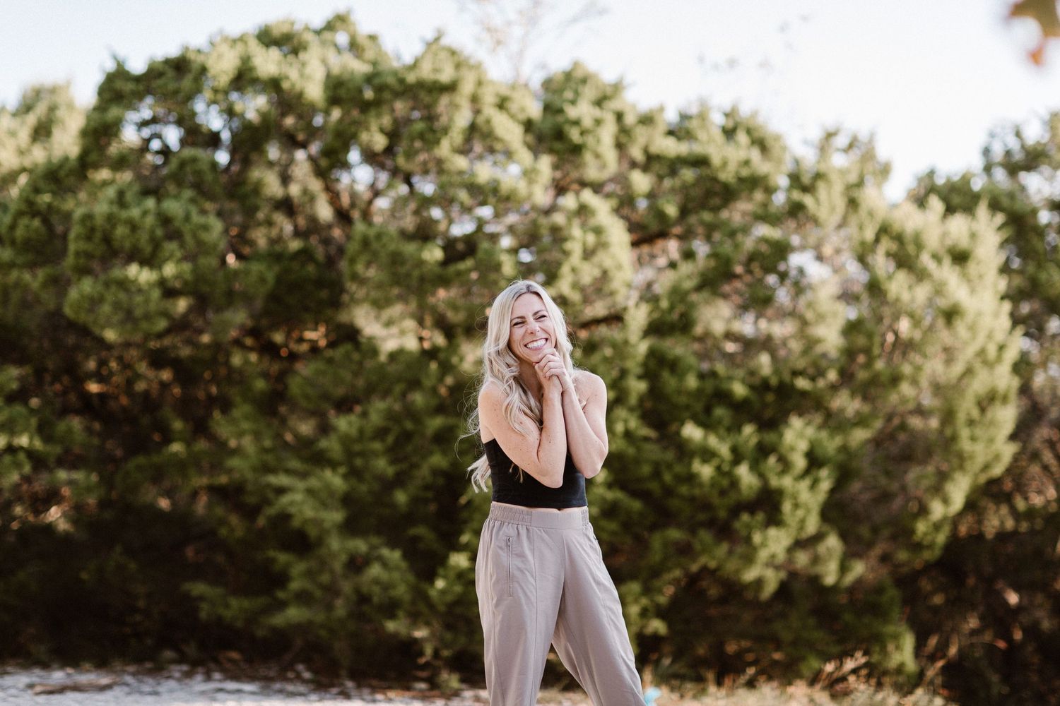 Blonde woman in black top and beige pants standing outdoors near lush green trees in natural light.