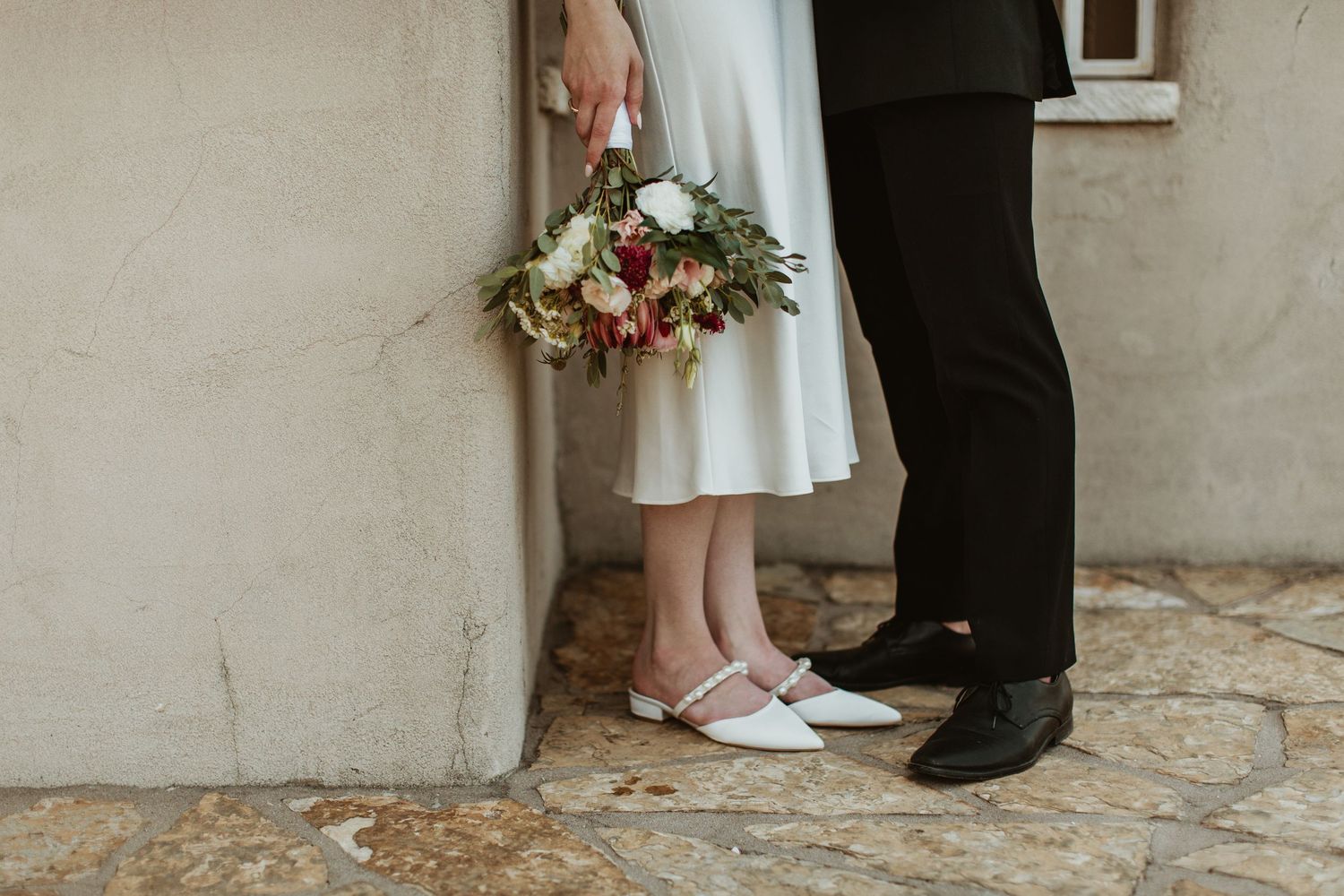 Bride and groom stand together, she holds a red and white bouquet, wearing white heels beside his black dress shoes.
