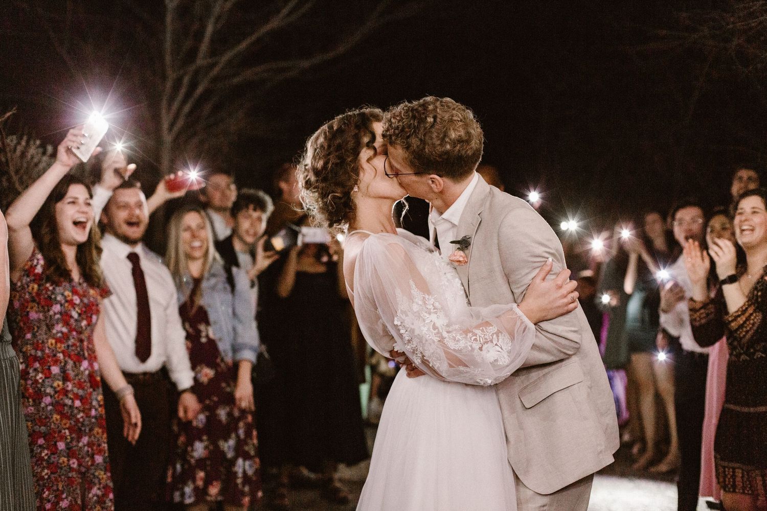 Bride and groom share first kiss surrounded by guests holding sparklers at romantic outdoor night wedding.
