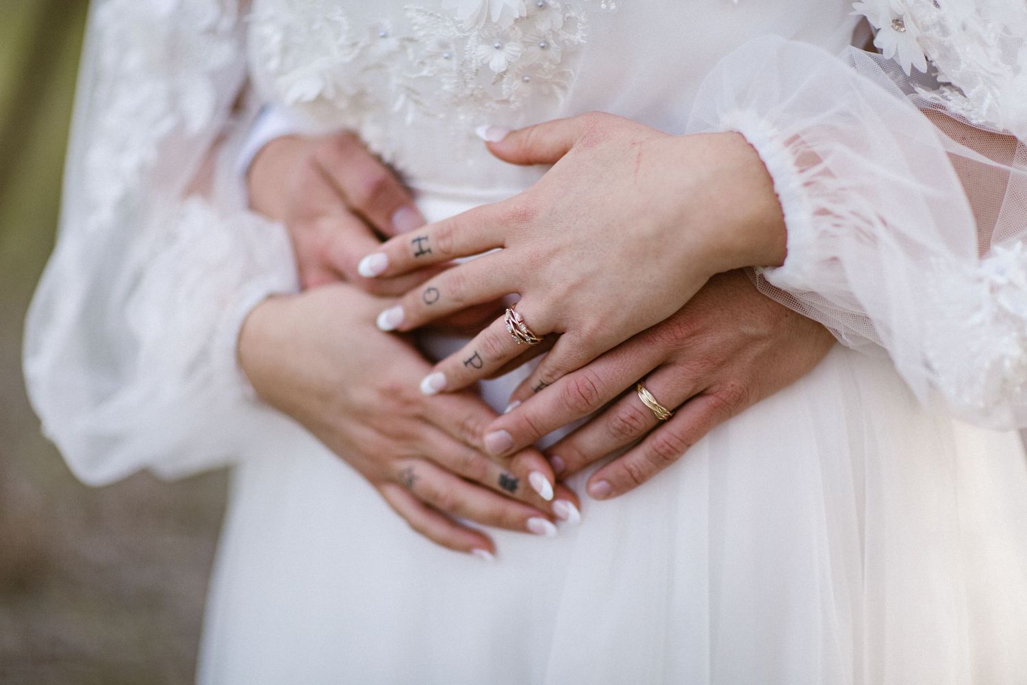 Close-up of bride and groom's hands with wedding rings clasped together over white bridal gown.