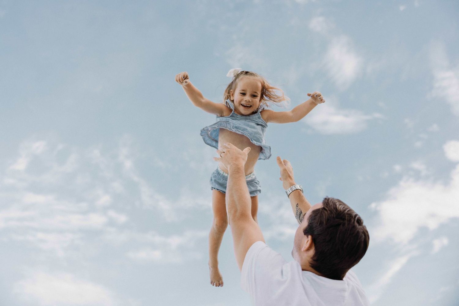 Father tossing his laughing young daughter high into the air against a bright blue sky.
