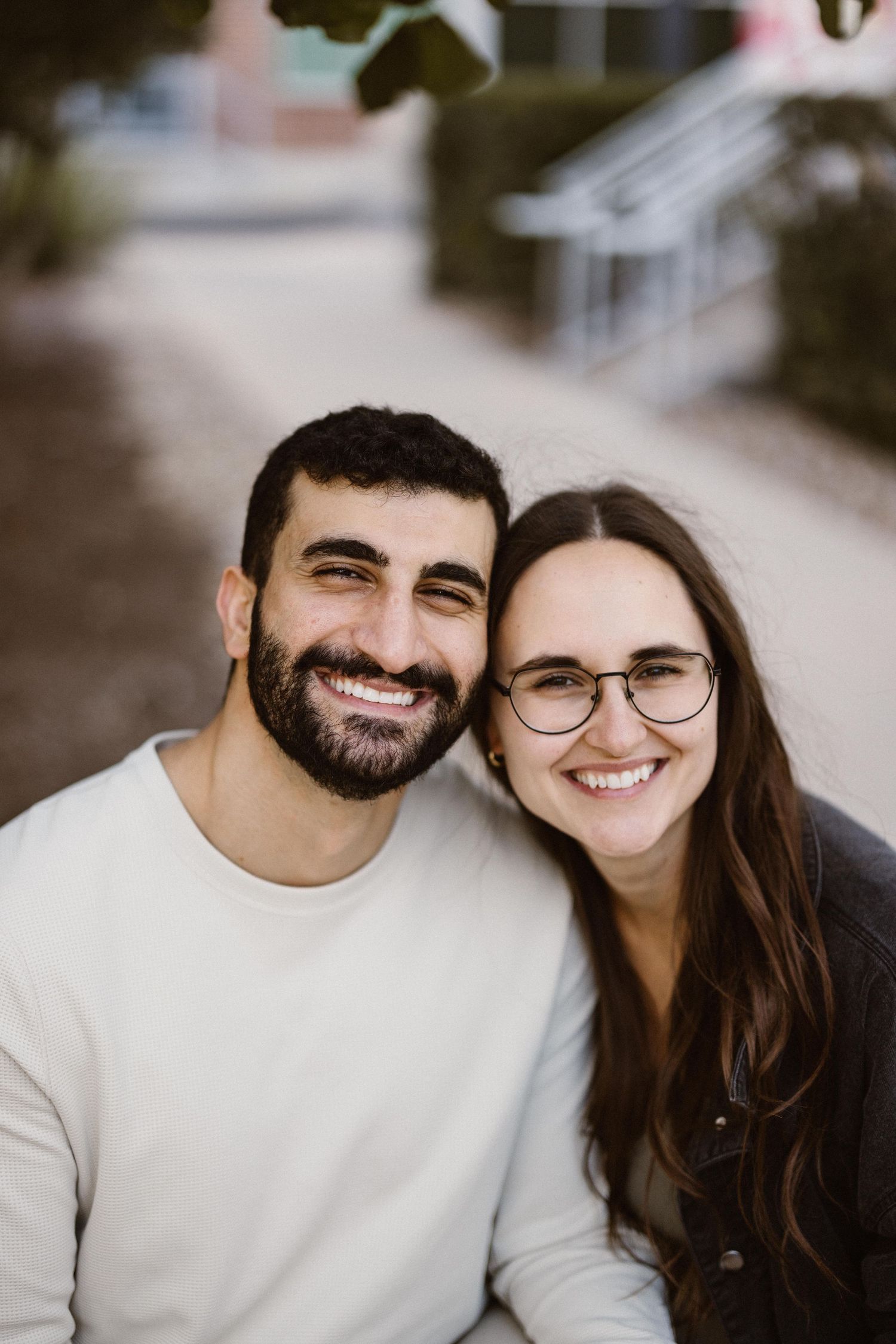 Happy couple smiling together outdoors, man in white sweater and woman in dark jacket wearing glasses.