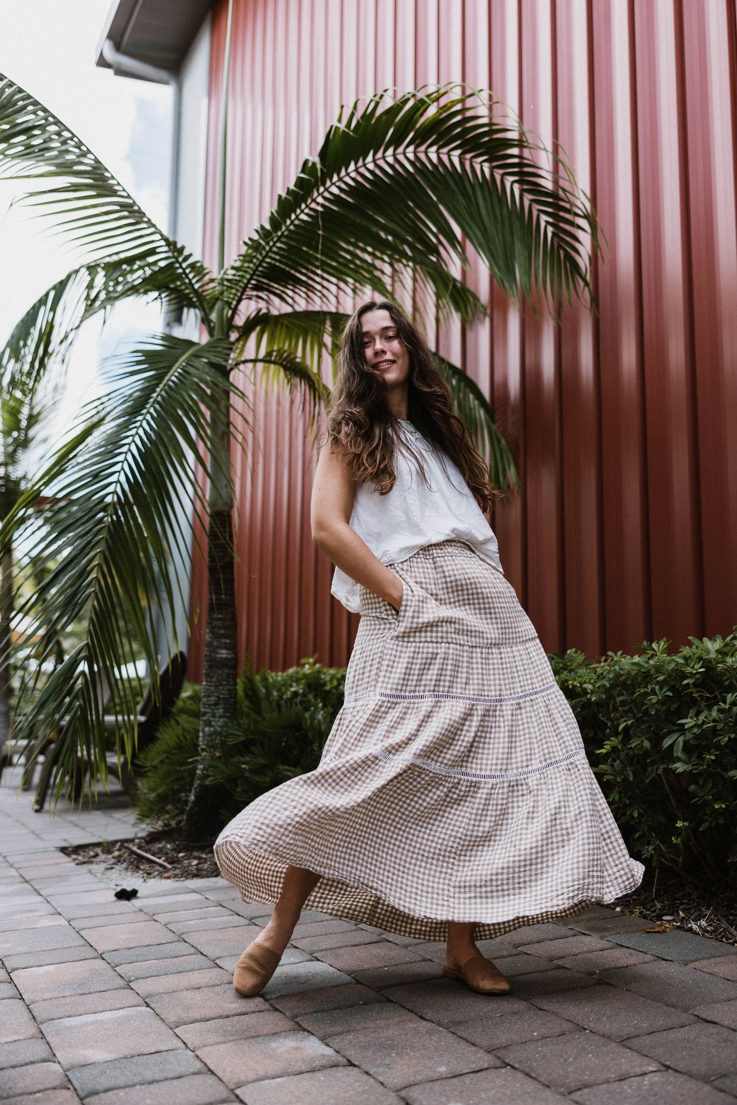 Young woman twirling in a plaid tiered maxi skirt near a palm tree against a red building.