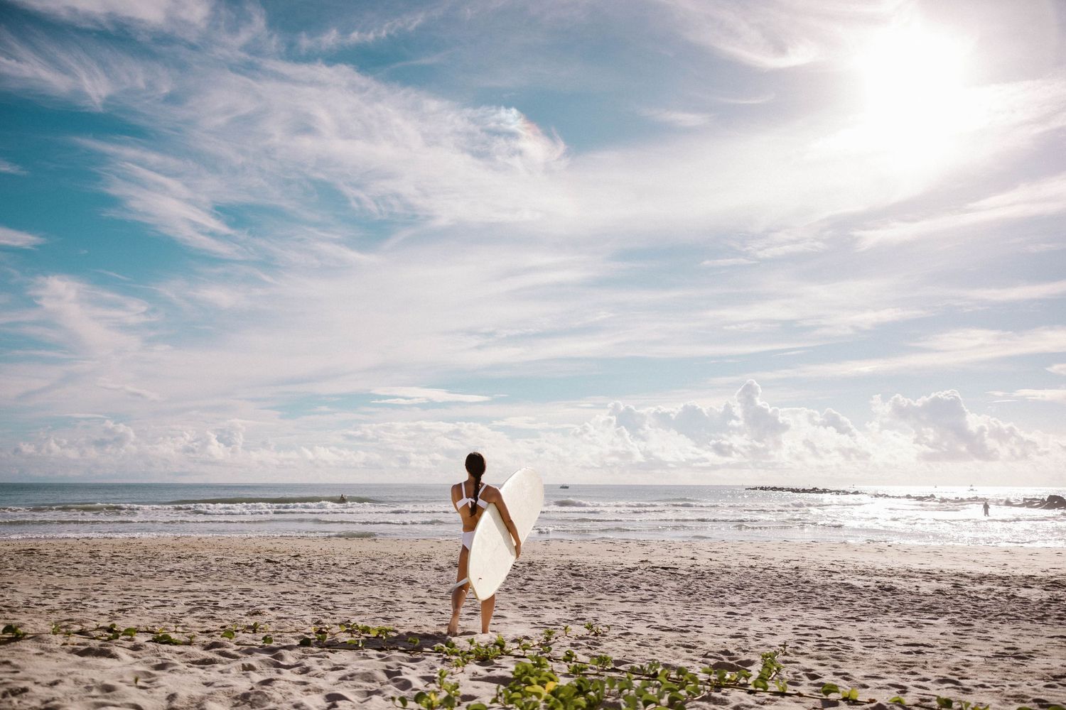 Woman carrying white surfboard walking on wide sandy beach under dramatic cloudy sky.