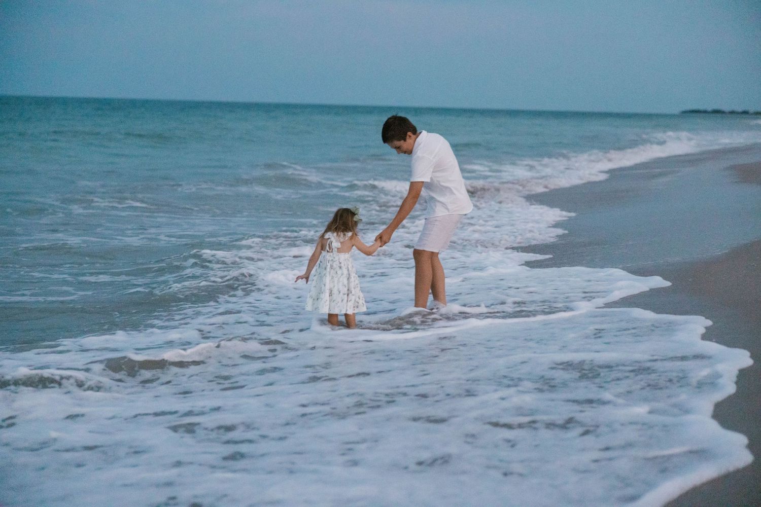 Father and young daughter holding hands while wading in ocean waves on beach.