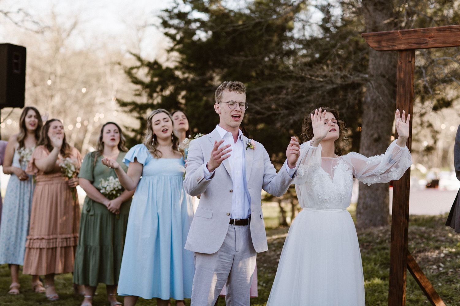 Bride and groom worship with hands raised during outdoor wedding ceremony while guests sing along behind them.