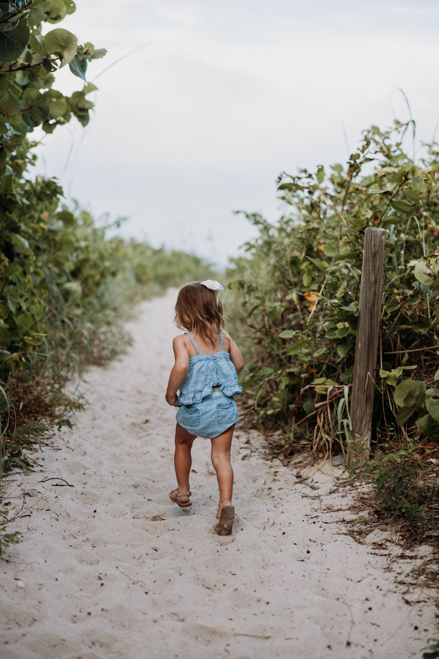 Young toddler girl in blue romper walking along a sandy beach path lined with tropical greenery.