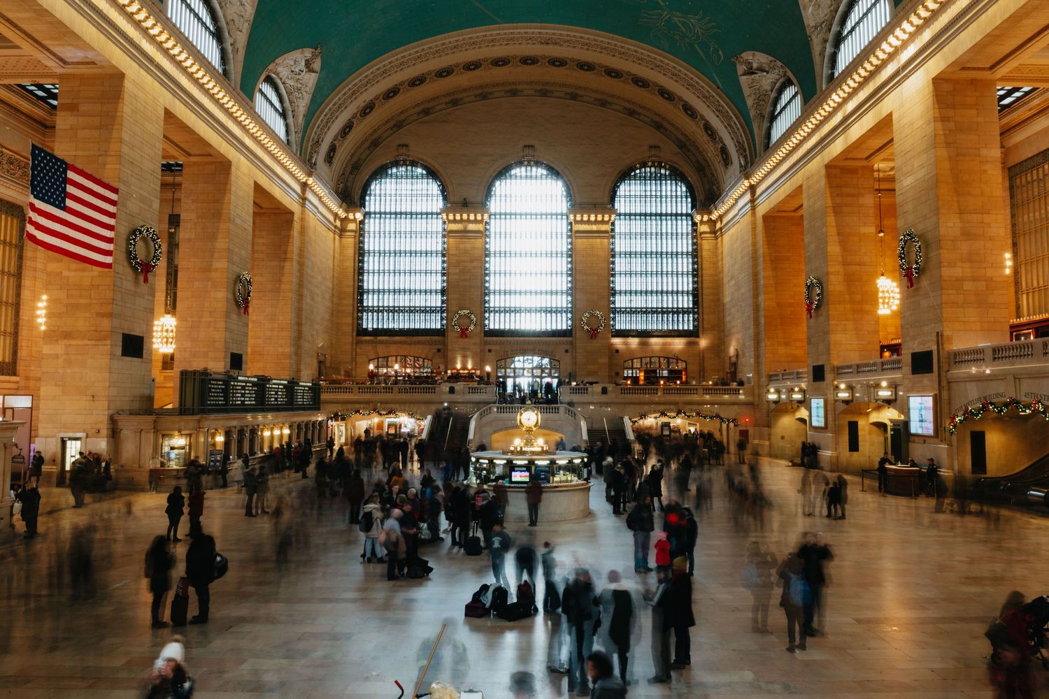 Busy Grand Central Terminal main concourse with crowds, arched windows, and American flag.