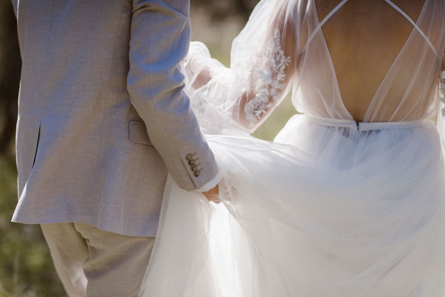 Close-up of bride in backless lace wedding gown holding grooms hand during outdoor ceremony.