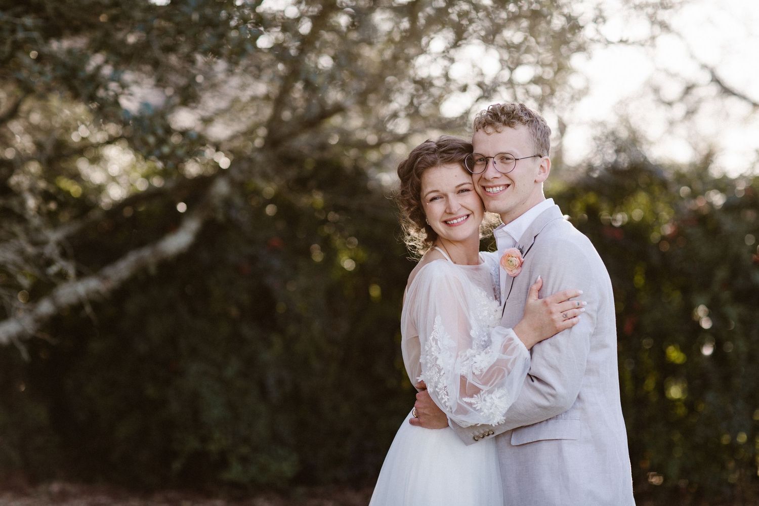 Smiling bride and groom embracing warmly in golden hour light with lush green trees behind them.
