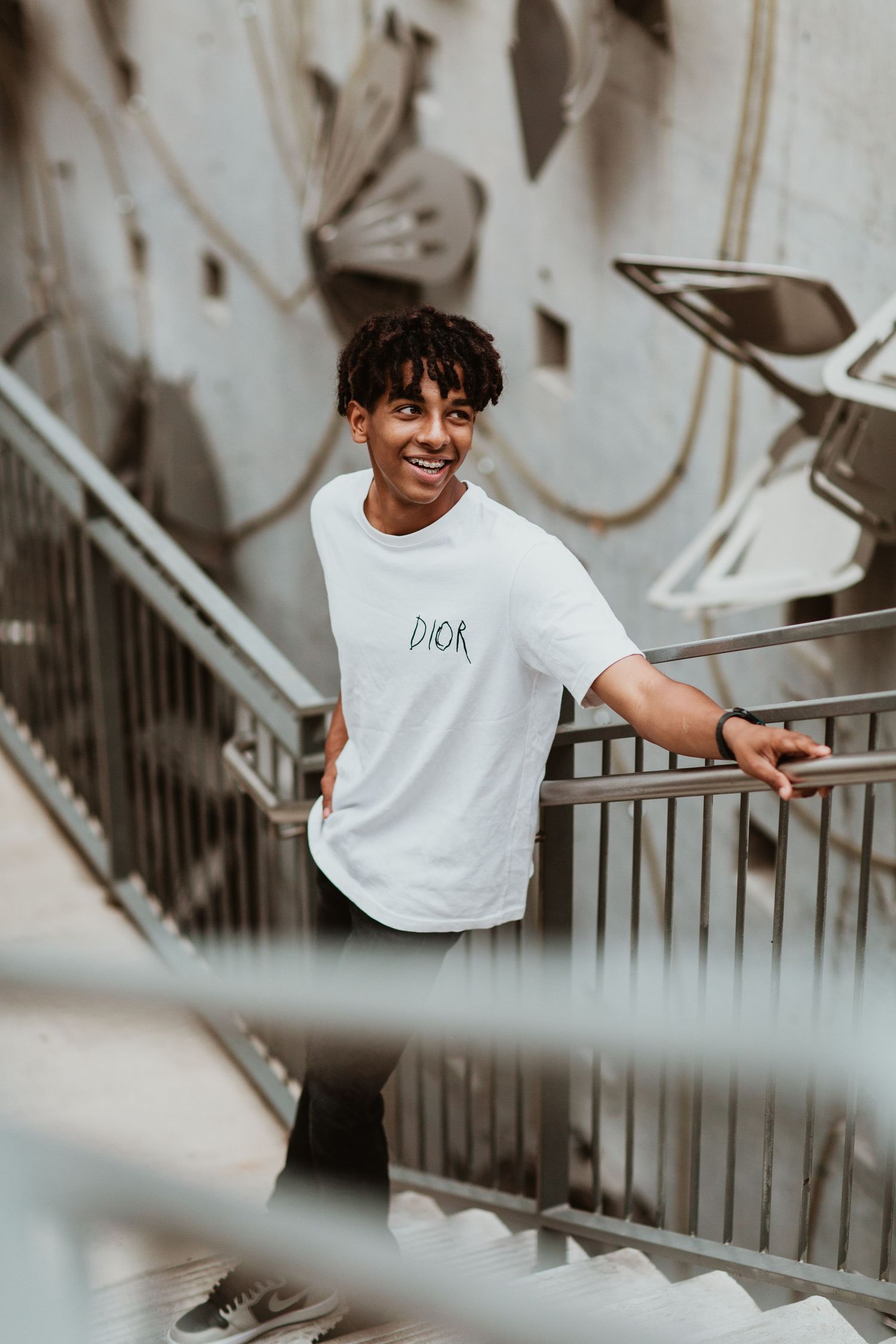Young man in white Dior t-shirt smiling on urban stairs with graffiti wall background.