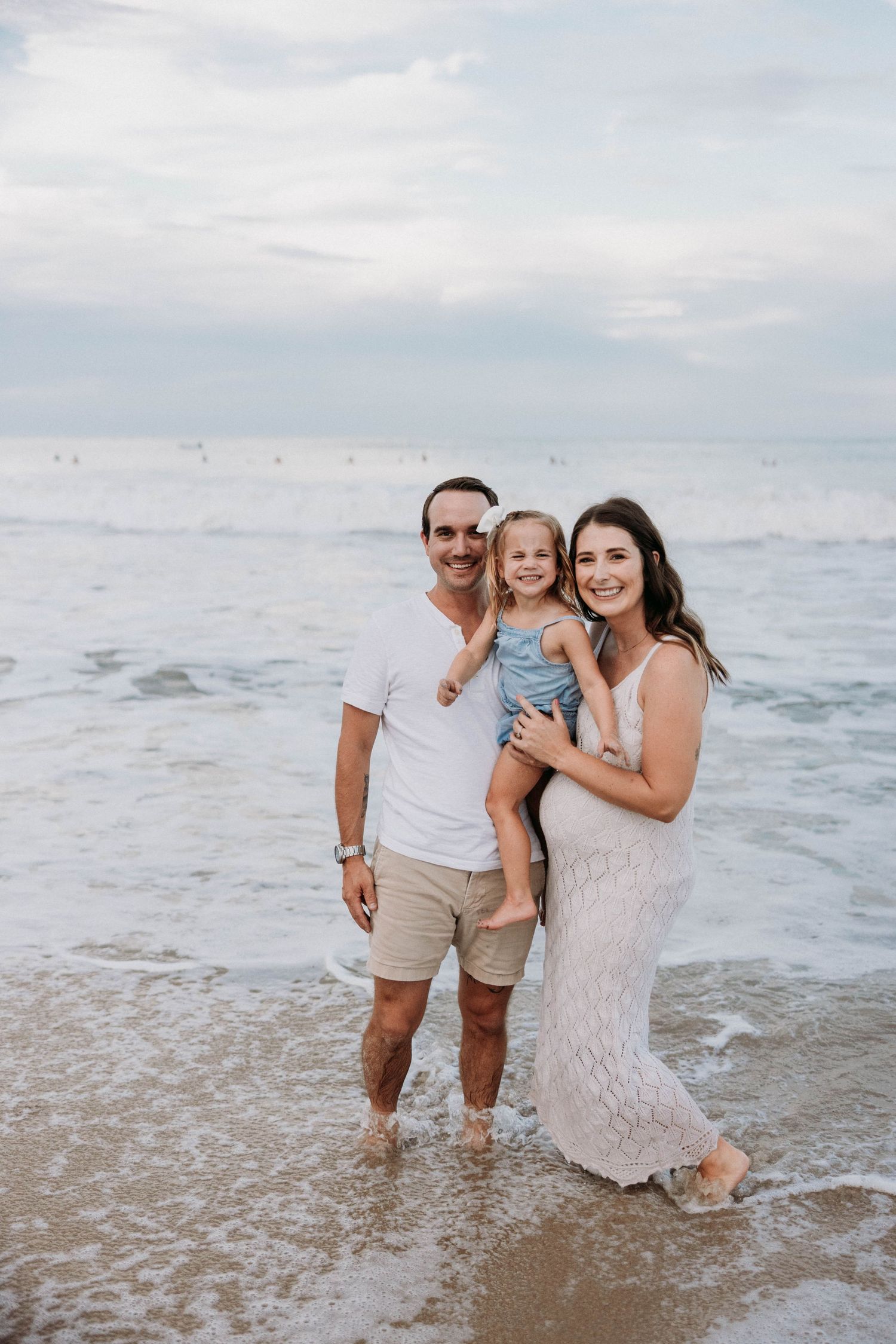 Happy family of three standing in ocean waves, father and pregnant mother holding young daughter.