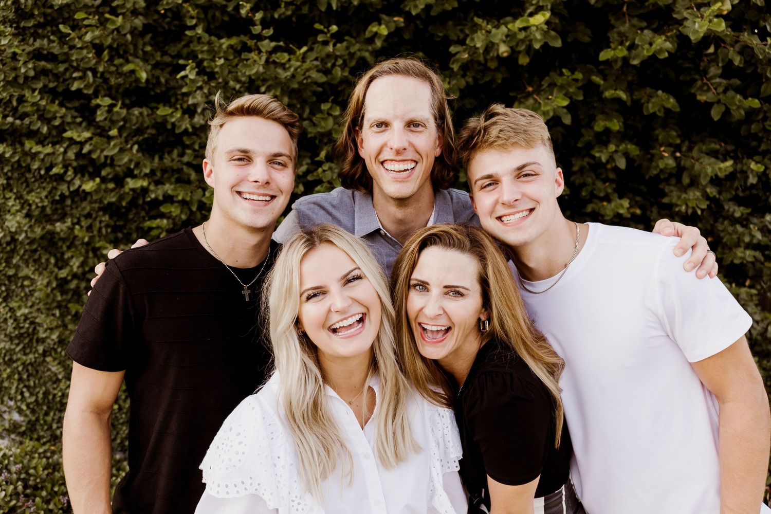 Happy family of five smiling together outdoors in front of a lush green hedge.