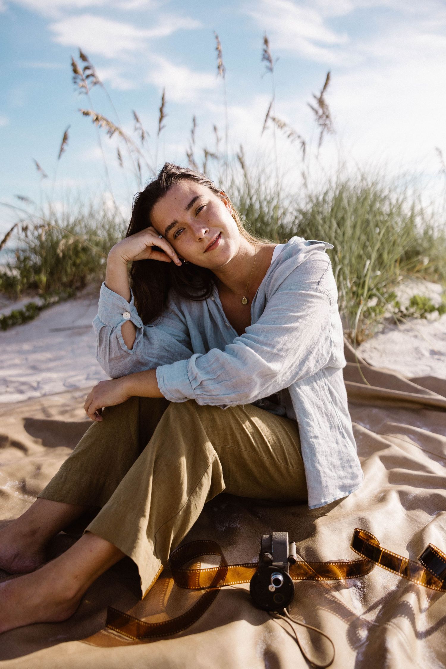 Woman in linen outfit sitting on sandy beach dunes with vintage camera, relaxing outdoors.