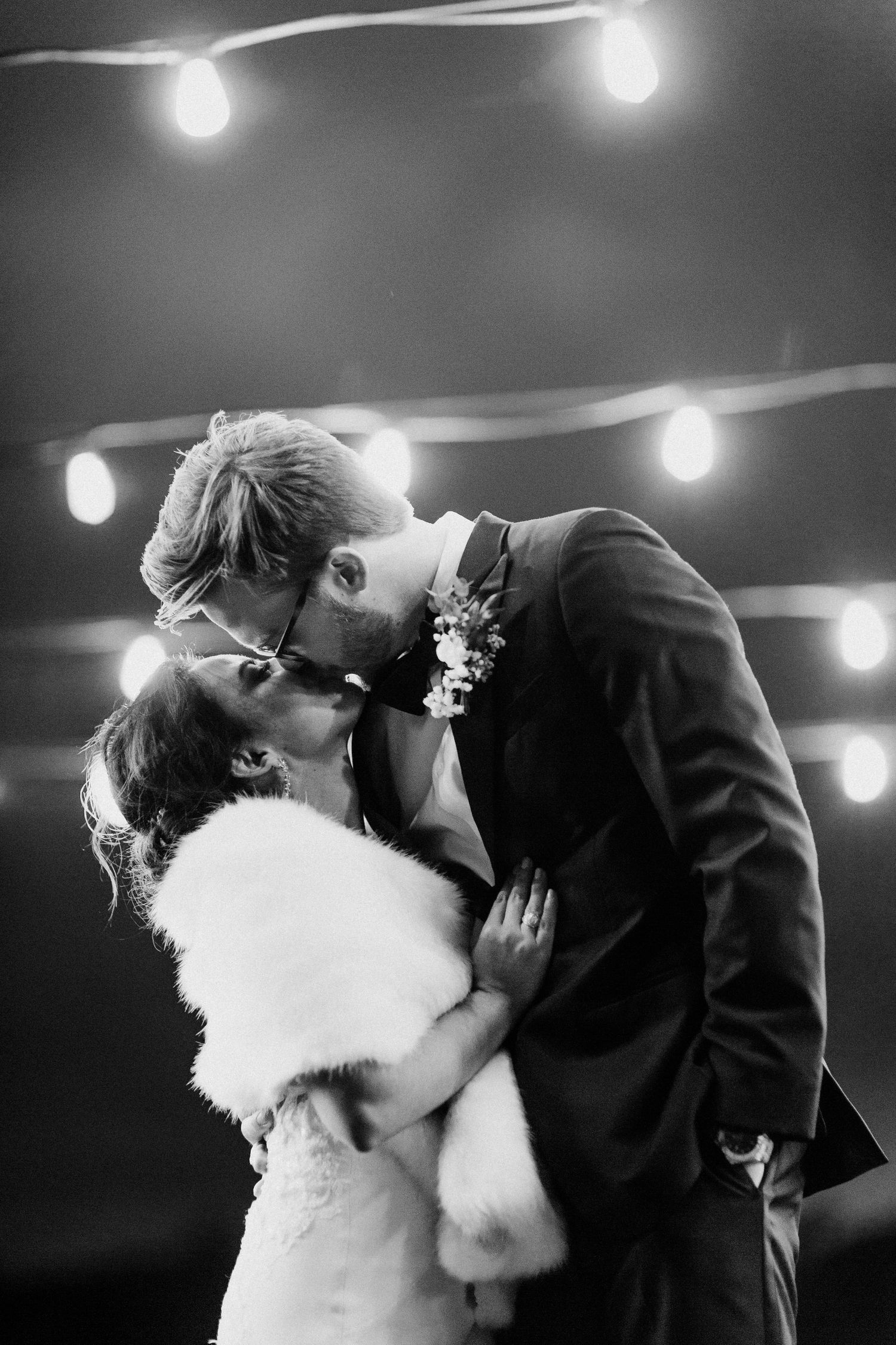 Black and white romantic photo of newlyweds sharing first kiss under string lights at wedding reception.