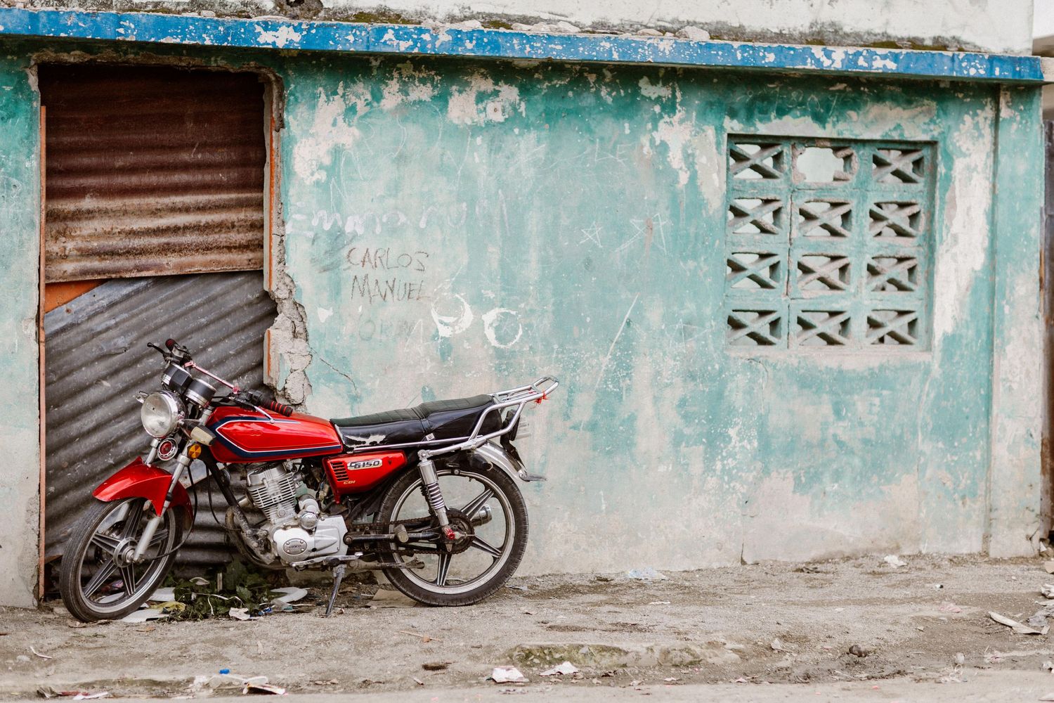 A red motorcycle parked against a weathered turquoise wall with a rusty metal door.
