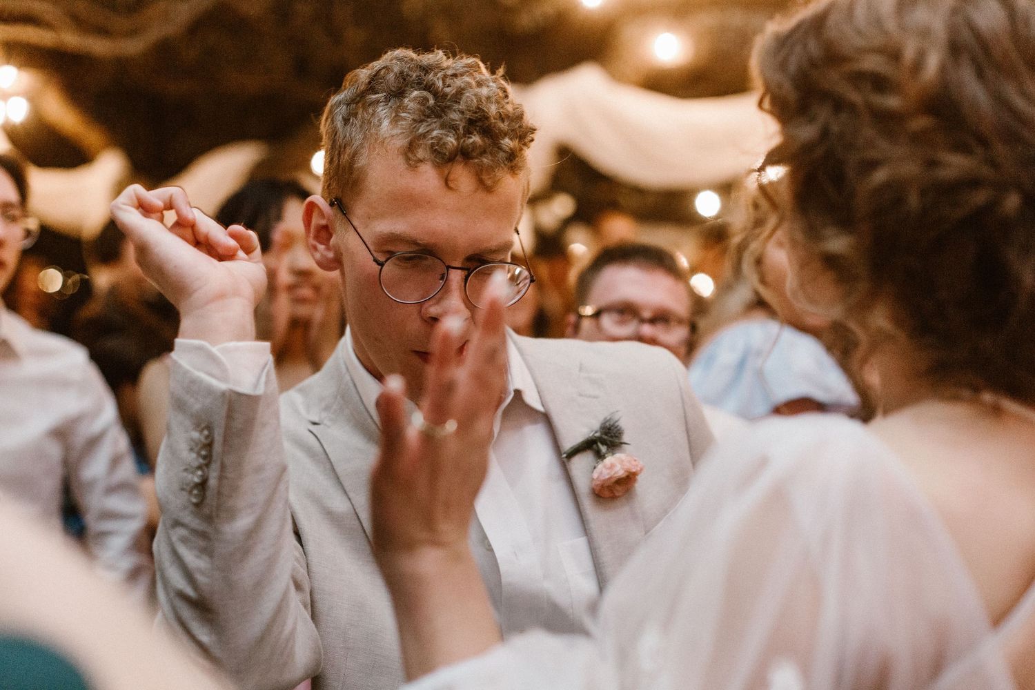 Groom in beige suit dancing emotionally at wedding reception with string lights.