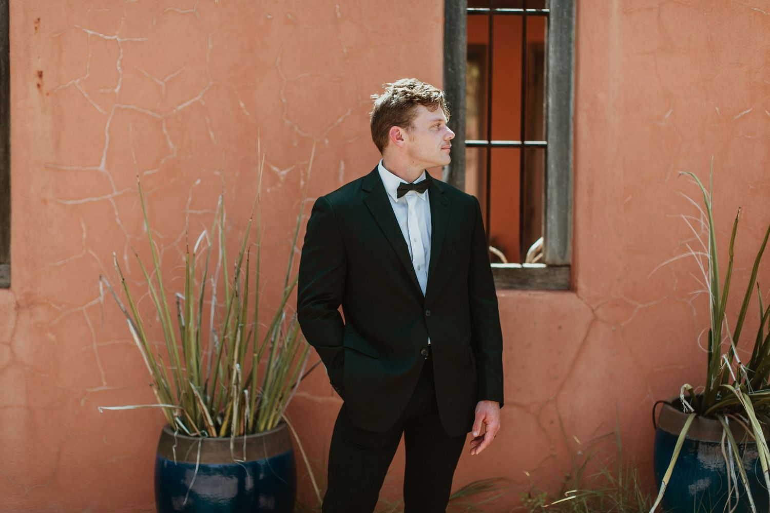 Groom in black tuxedo and bow tie standing against a terracotta wall with potted plants.