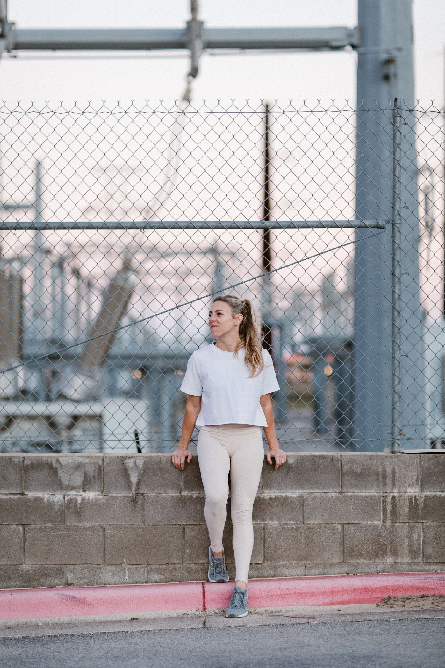 Woman in white crop top and leggings leaning against a concrete wall with chain-link fence in urban setting.