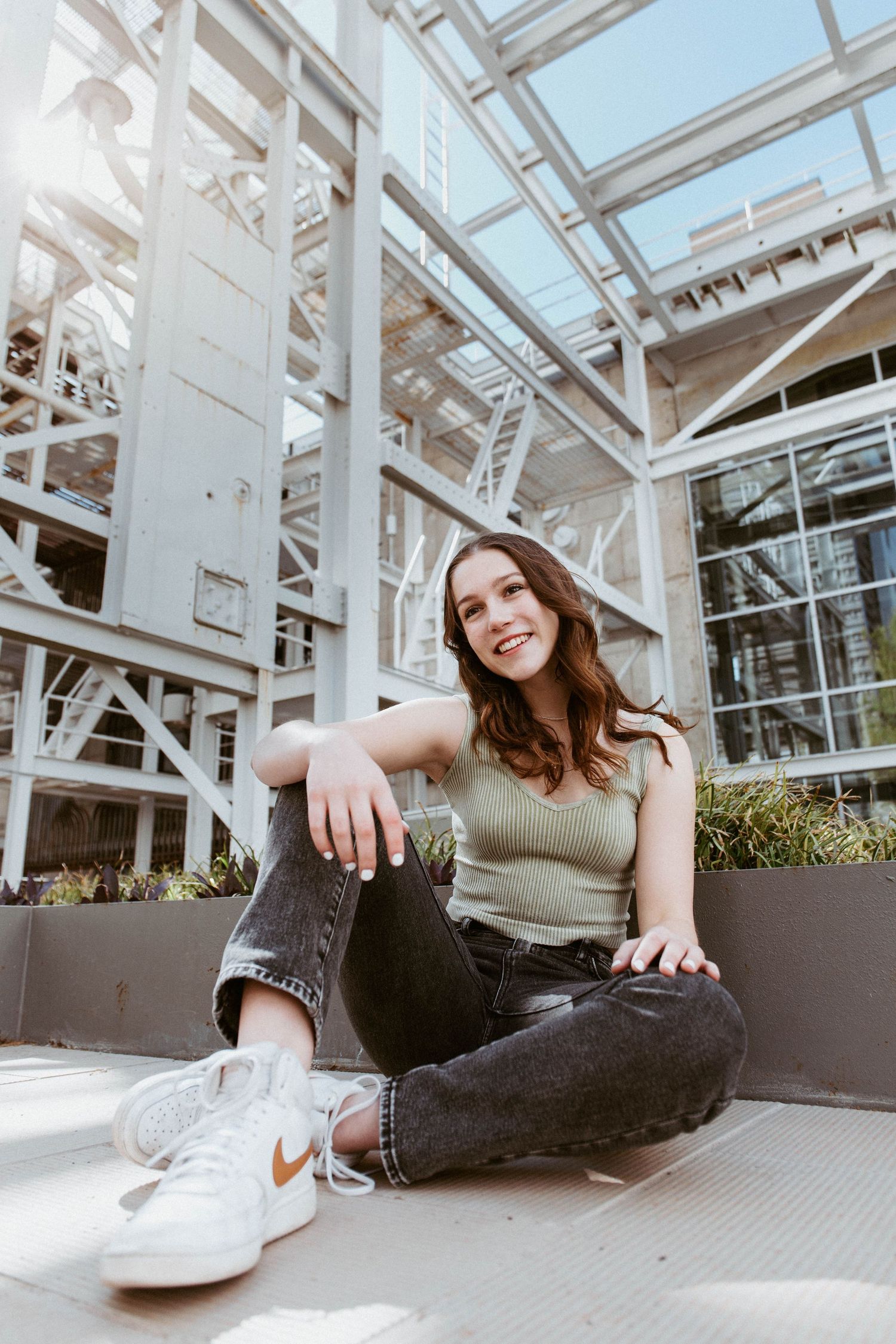 Smiling young woman in jeans and Nike sneakers sits relaxed beneath a modern glass structure outdoors.