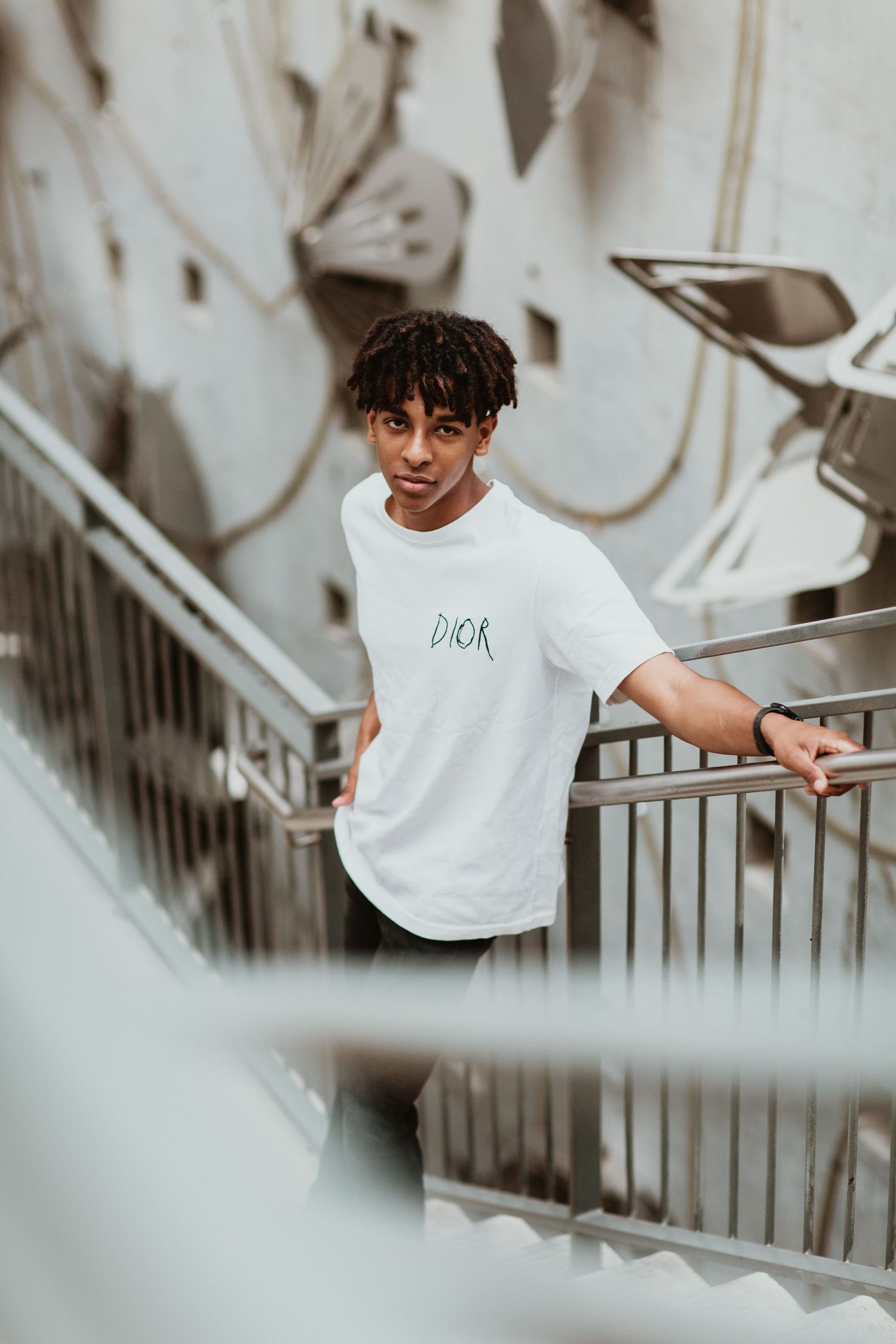 Young man in white Dior t-shirt leaning on metal staircase railing with graffiti wall background.
