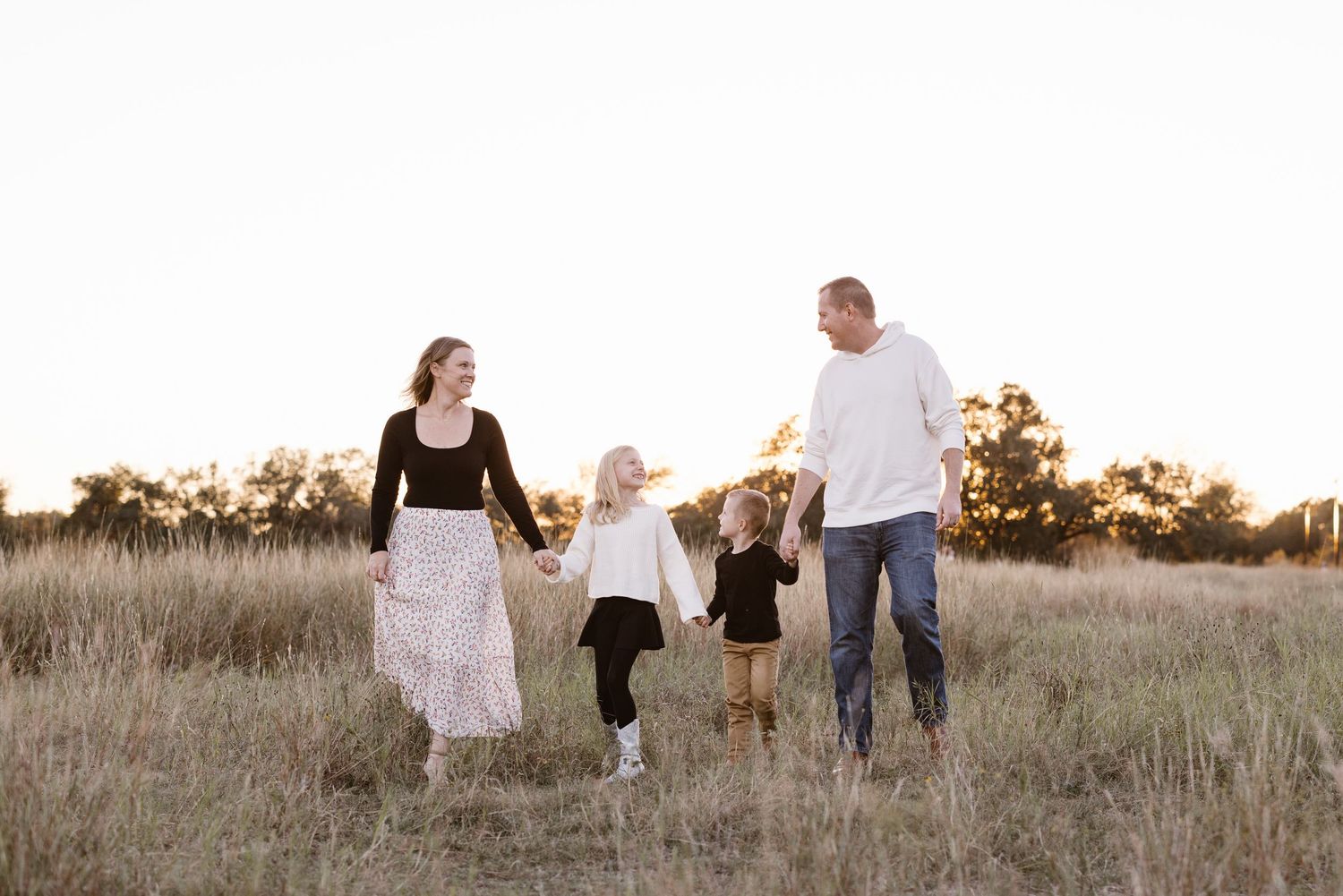 Happy family of four holding hands walking through a golden field at sunset.