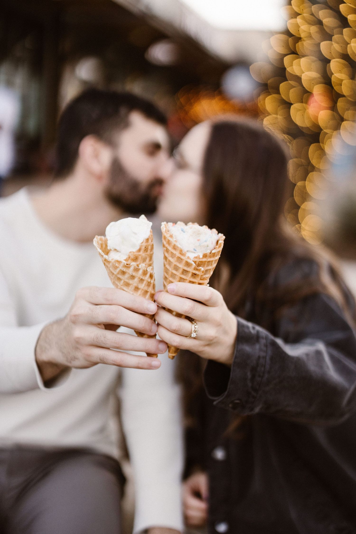 Romantic couple kissing while holding two waffle ice cream cones with bokeh lights.