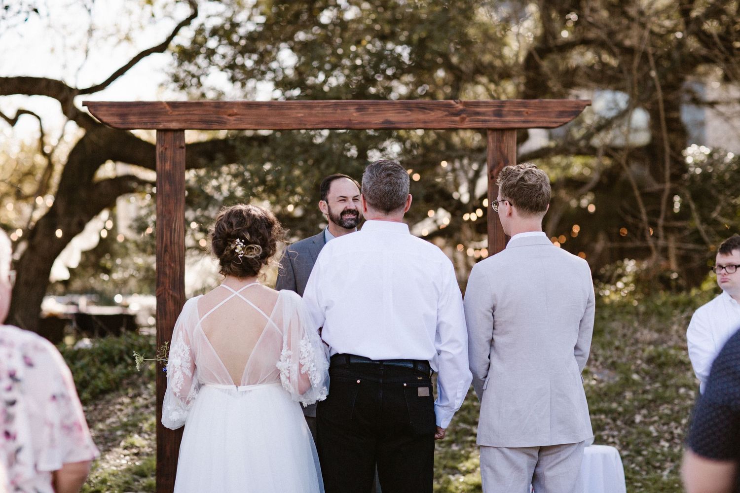 Bride and groom stand before the officiant under a rustic wooden arch surrounded by guests at an outdoor wedding ceremony.