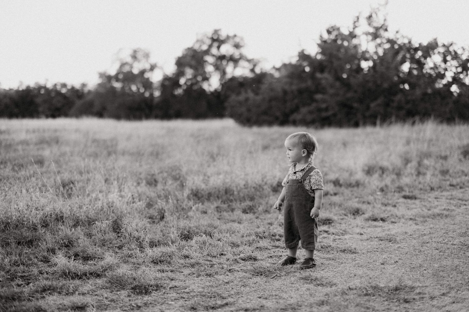 A toddler stands alone on a dirt path in a grassy field in a black and white photo.