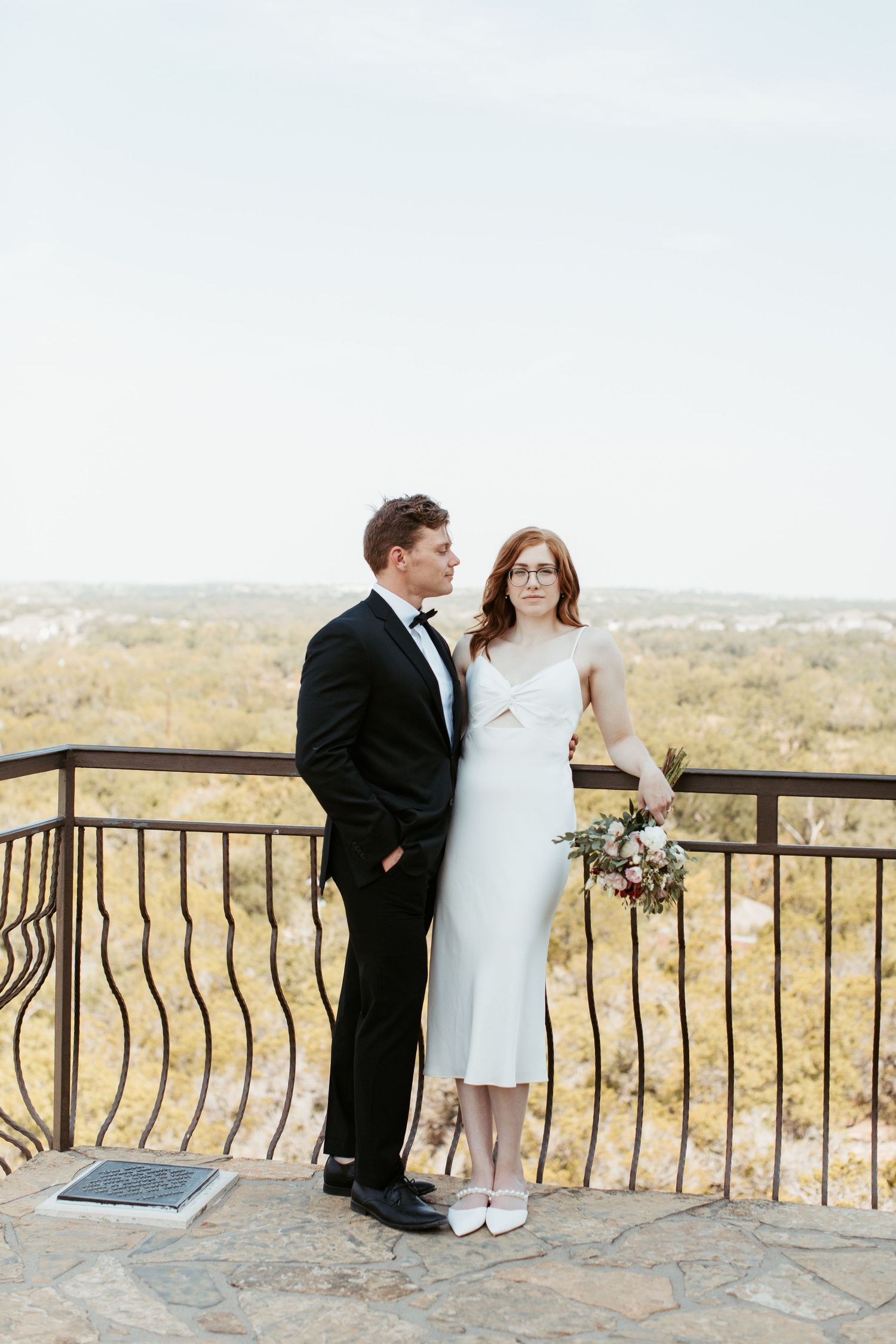 Bride and groom pose on scenic overlook balcony, she in white midi dress with bouquet, he in classic black tuxedo.