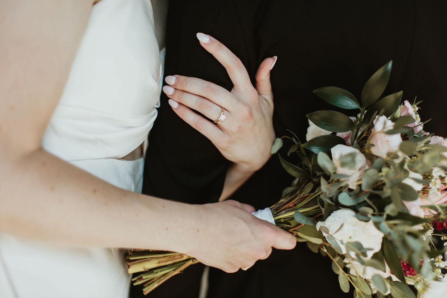 Bride holding a floral bouquet with engagement ring visible, standing next to groom in black suit.