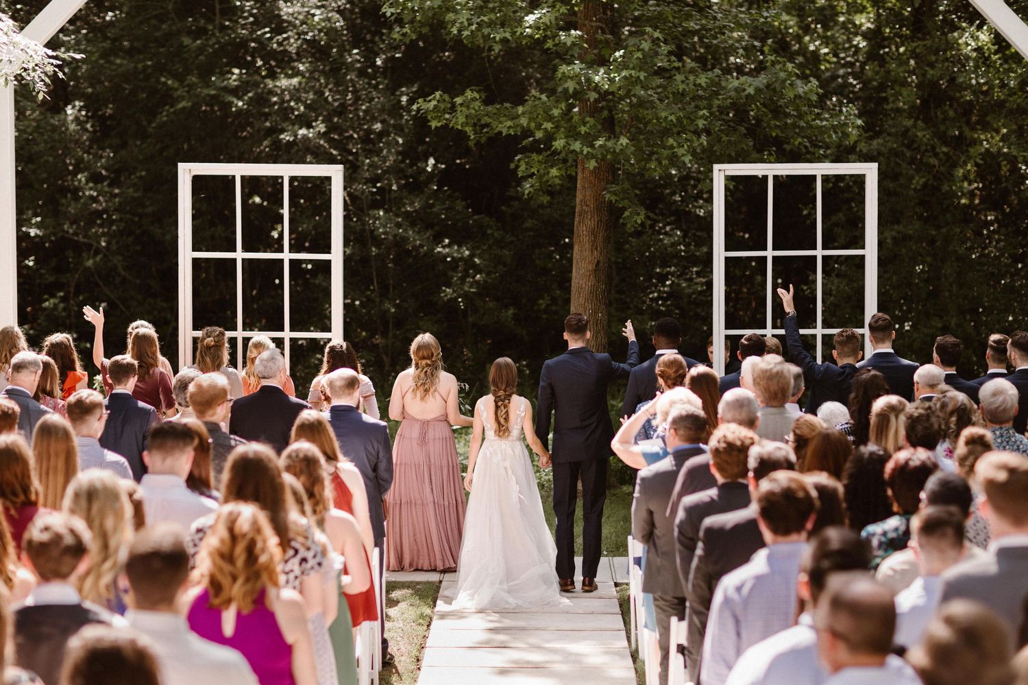 Bride walking down outdoor aisle toward altar surrounded by standing wedding guests.