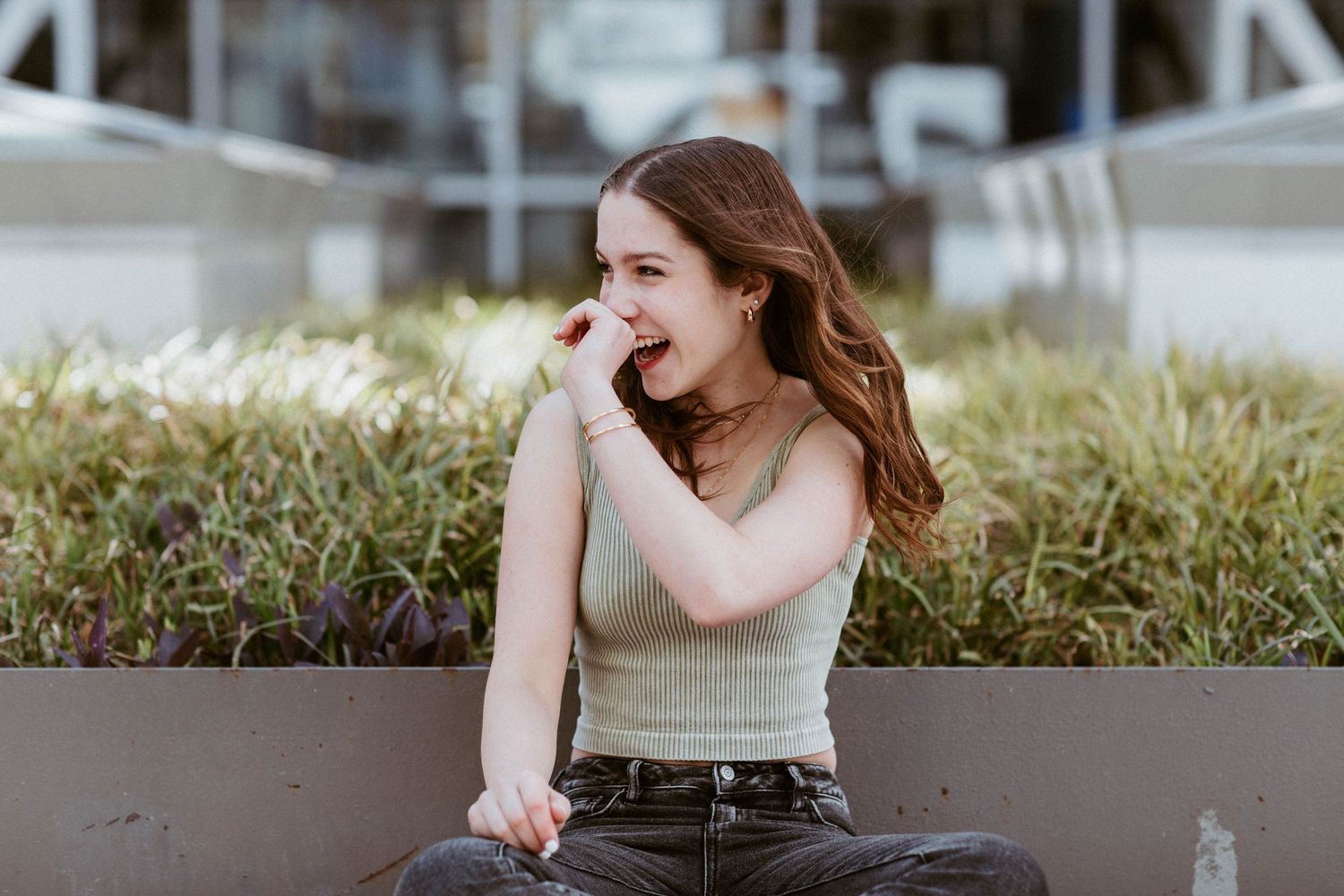 Cheerful young woman in a green crop top laughs while sitting cross-legged outdoors near greenery.