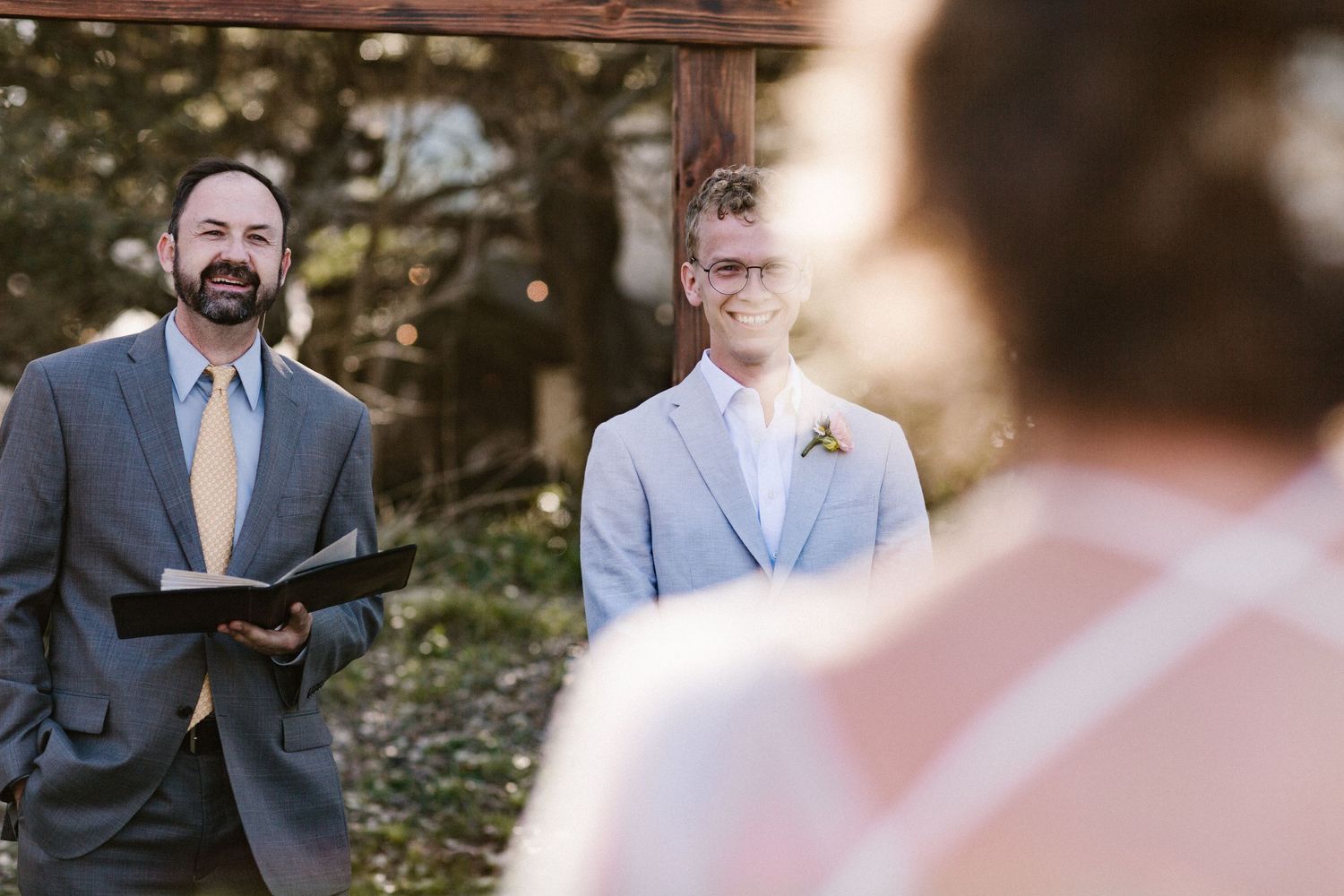Groom in light gray suit stands at the altar smiling as the officiant in a gray suit reads from a book at the ceremony.